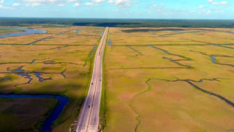 Aerial view of a two-lane highway crossing expansive South Carolina salt marsh, with golden-green cordgrass divided by winding tidal creeks, a tree-lined shoreline on the horizon, and a bright blue sky with scattered cumulus clouds.